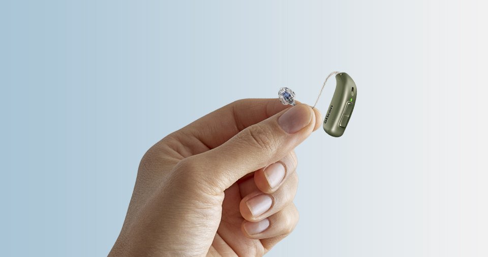 Hand holding a small hearing aid with a clear earpiece, set against a gradient blue to white background. The hearing aid has a visible button and green indicator light.