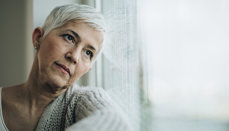 Woman with short gray hair gazing out of a rain-streaked window while resting on her arm.