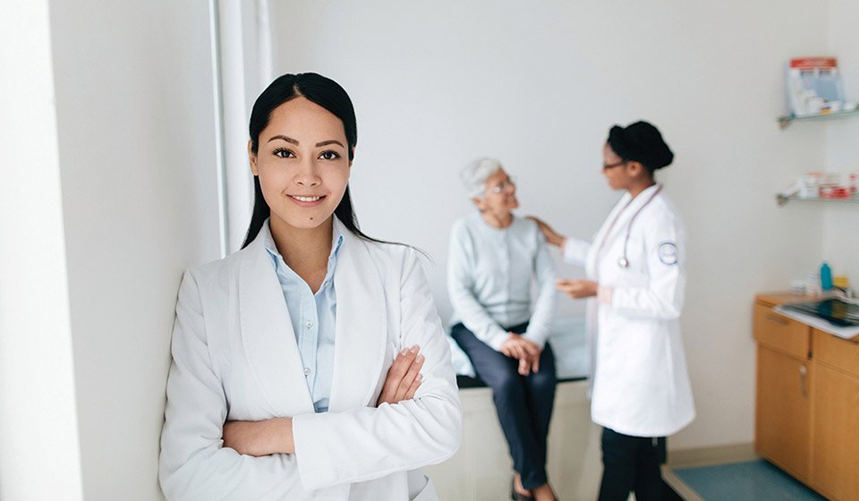 Audiologist standing confidently in a hearing clinic while a patient consults with another healthcare professional.