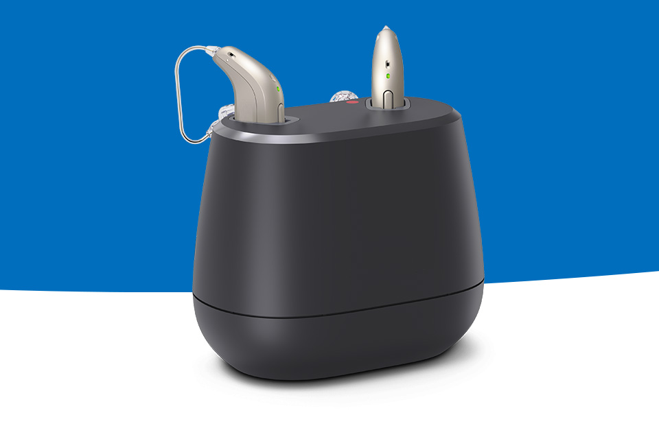 Rechargeable hearing aids in a modern black charging station against a blue and white background.