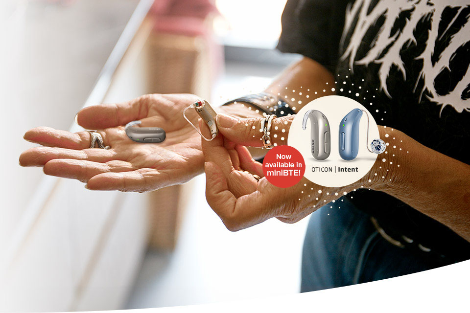 Hands holding hearing aids; background shows a bright indoor setting. A circular inset depicts two new hearing aids with text: OTICON Intent - Now available in miniBTE!