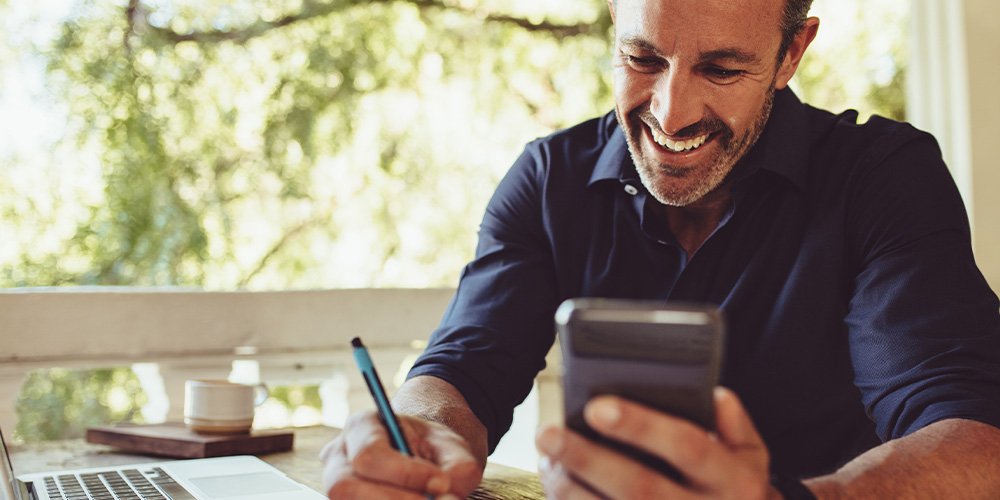 Man in navy shirt holding a smartphone and taking notes with a pen while sitting at a desk with a laptop and mug.