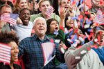 People enthusiastically wave small American flags while cheering in a crowd, surrounded by others expressing joy and excitement in a lively, celebratory environment.