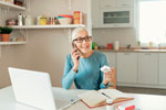 An older woman talks on the phone while holding medication, seated at a kitchen table with a laptop, notebook, and eyeglasses. The kitchen has shelves with various items and a window.