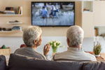 Two elderly individuals, seated on a couch, are watching television in a modern and neatly organized living room with shelves, books, and a vase of flowers.