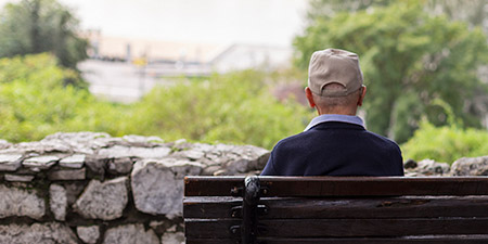 Man wearing a beige cap sitting alone on a wooden bench in a park, overlooking greenery and a stone wall.