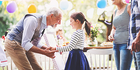 Grandfather giving a young girl a birthday present at an outdoor party while family gathers nearby.