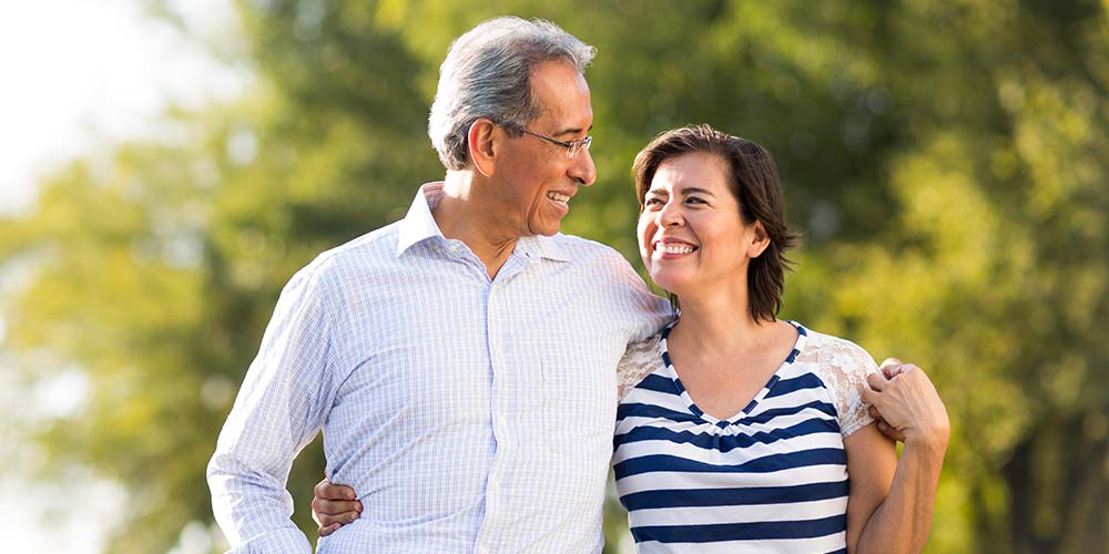 A smiling man and woman walk arm-in-arm outdoors, exchanging happy glances, surrounded by lush green trees in a sunny setting.