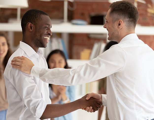 Two smiling men in white shirts shake hands warmly in a modern office, with colleagues in the background looking on and clapping. Shelves and brick walls are visible in the background.