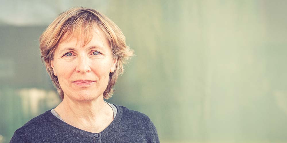 A woman, with short blonde hair and wearing a dark sweater, looks directly at the camera in front of a blurred, light-colored background.