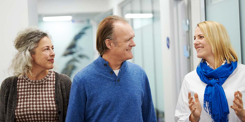 Three people converse while walking down a hallway; two women and a man are smiling, dressed in casual business attire, the hallway features glass walls and bright lighting.