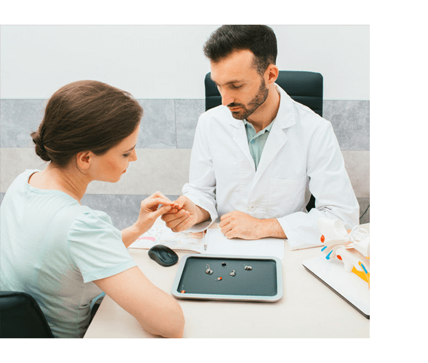 A doctor in a white coat shows a hearing aid to a seated woman in an office. The table has various hearing aids, a mouse, and papers.