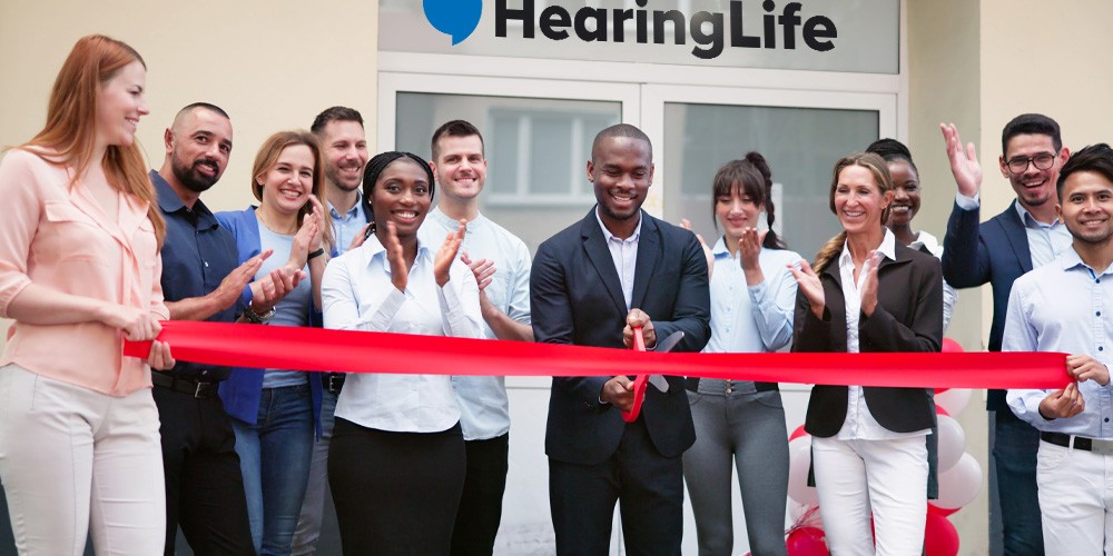 Grand opening celebration at a HearingLife center with a ribbon-cutting ceremony, featuring a diverse group of people smiling and clapping.