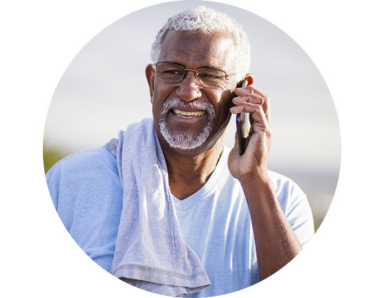 Man with gray hair smiling while talking on a mobile phone, wearing glasses, a light blue shirt, and a towel draped over his shoulder.