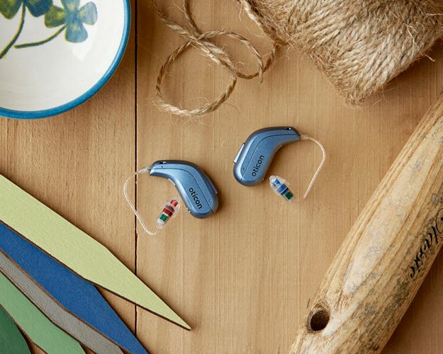 Behind-the-ear hearing aids placed on a wooden surface surrounded by crafting materials, including string, felt, and a paintbrush.