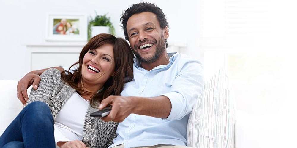 A man and woman are sitting on a couch, laughing, with the man holding a remote control. Behind them, there's a white shelf with a framed photo and a green plant.
