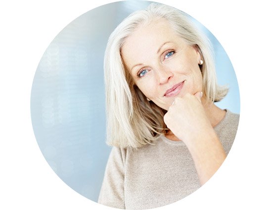 Smiling woman with short gray hair touching her chin, wearing a neutral-colored sweater against a soft blue background.