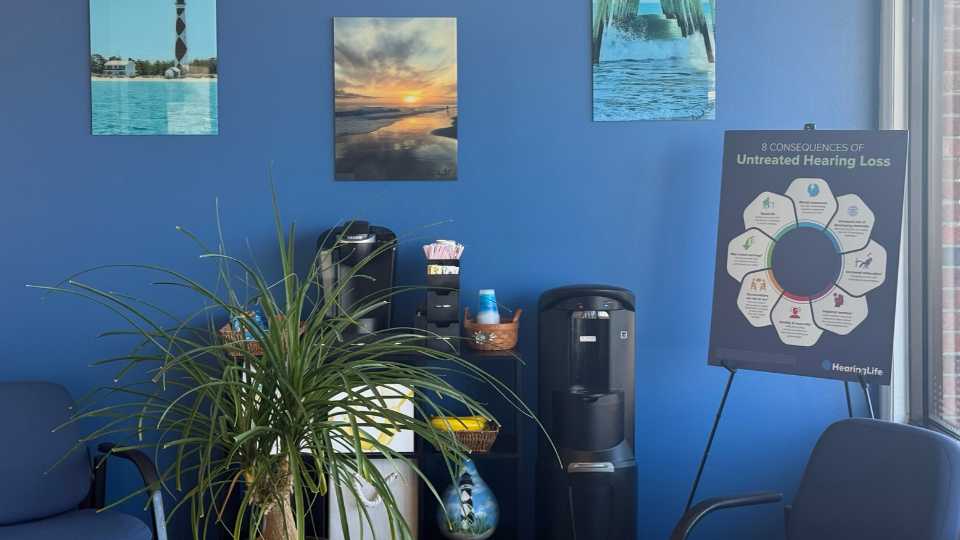 Hearing care clinic lobby in Cedar Point, North Carolina featuring blue walls, decorative artwork, informational poster on untreated hearing loss, seating area, and refreshment station.
