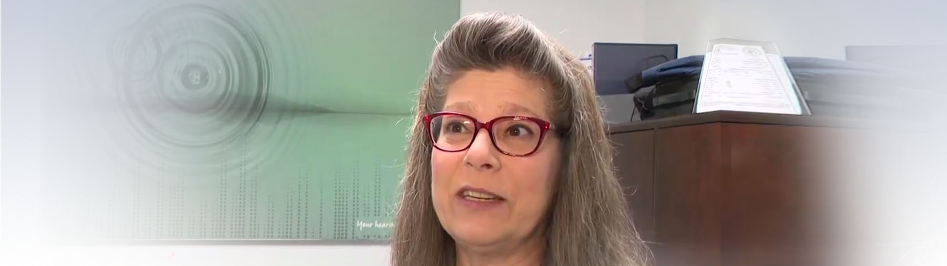 Hearing care professional in a clinic with a diploma and soundwave artwork displayed in the background.