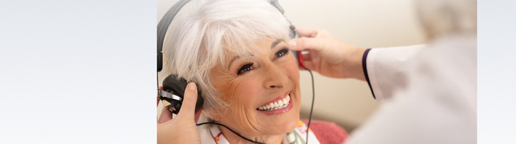 A smiling woman wears headphones as an audiologist adjusts them during a hearing test in a professional hearing clinic, indicating care and expertise in hearing health services.