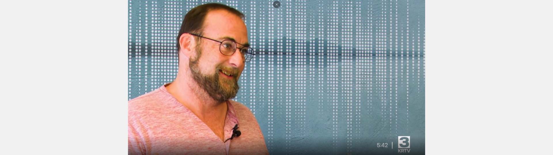 Smiling man in glasses and a pink shirt in front of a blue patterned background with vertical rows of white dots.