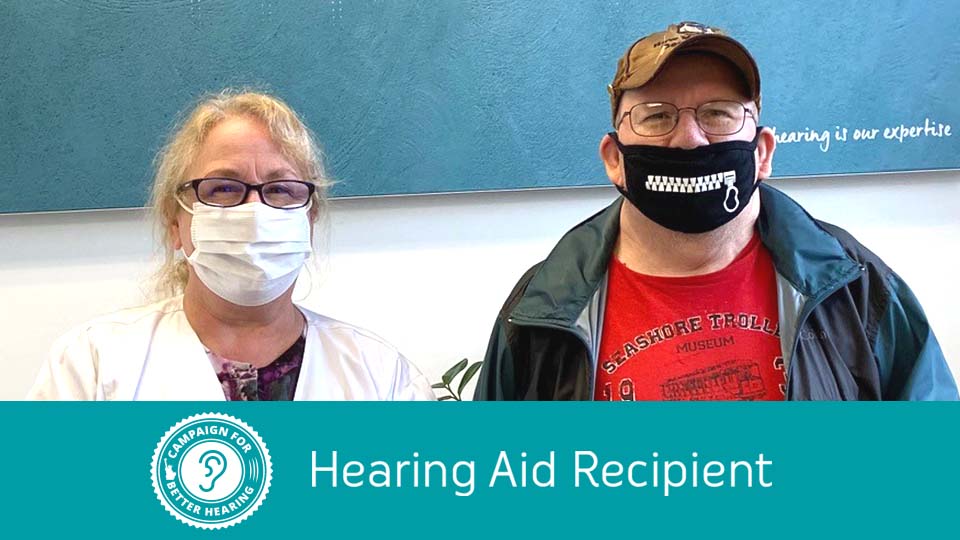 Two masked individuals, one in a white coat and one in casual attire, stand indoors beside a sign promoting the Campaign for Better Hearing. The sign reads: 'Hearing Aid Recipient – Hearing is our expertise.'