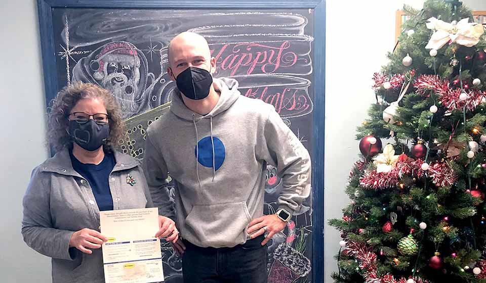 Two masked individuals indoors near a decorated Christmas tree; one person holds a certificate. A chalkboard in the background features a drawing of Santa Claus and the text 'Happy Holidays.'
