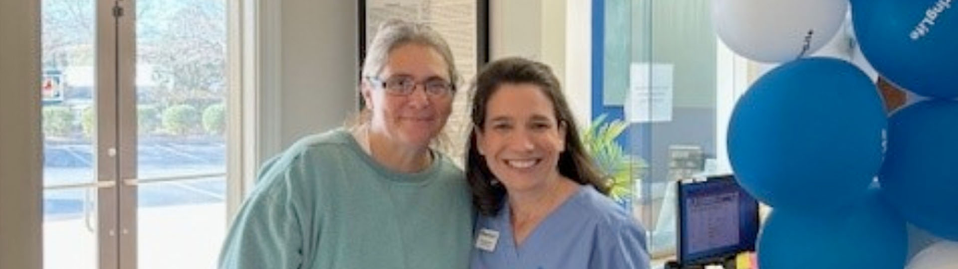 Kim Liscomb with a hearing care professional in a clinic near blue and white balloons.