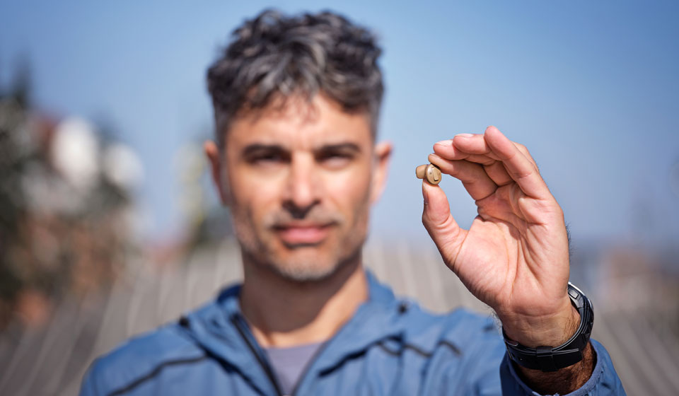 A man holds a small, beige hearing aid in his right hand while outdoors, wearing a blue jacket and a black wristwatch against a blurred background.