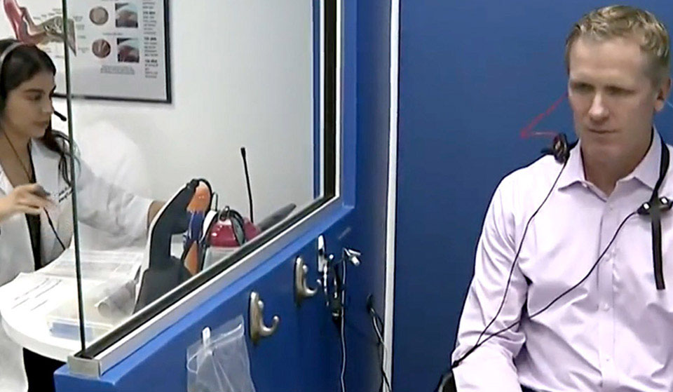 A man in a pink shirt with headphones sits in a blue-walled audiology booth, while a female audiologist, separated by a glass partition, operates equipment in a clinical setting.