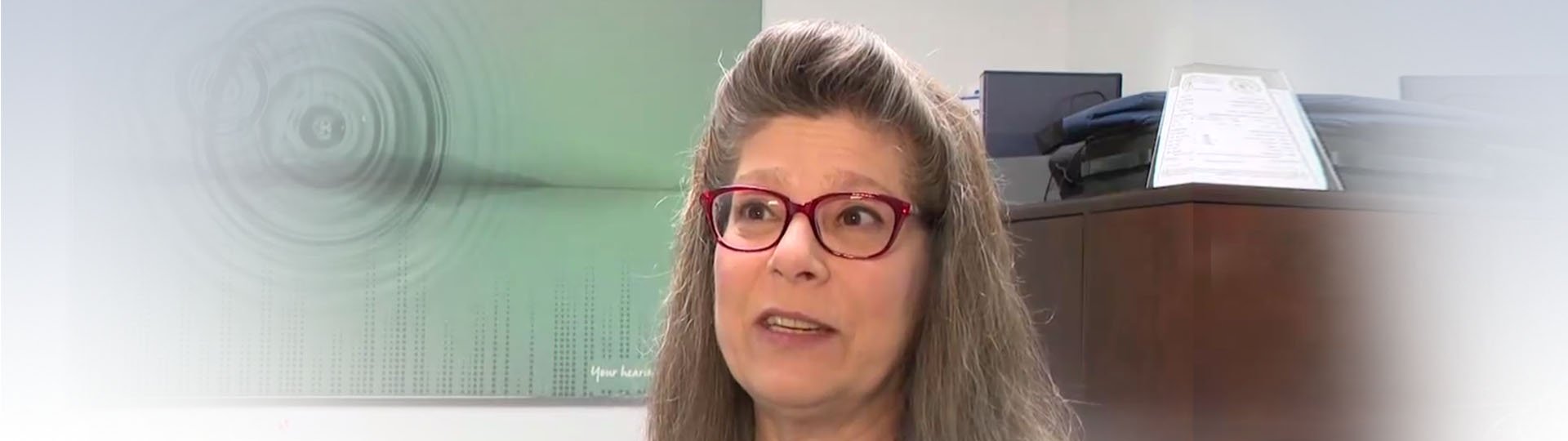 Adora Engstrom in a hearing care clinic setting with a diploma and soundwave artwork in the background.