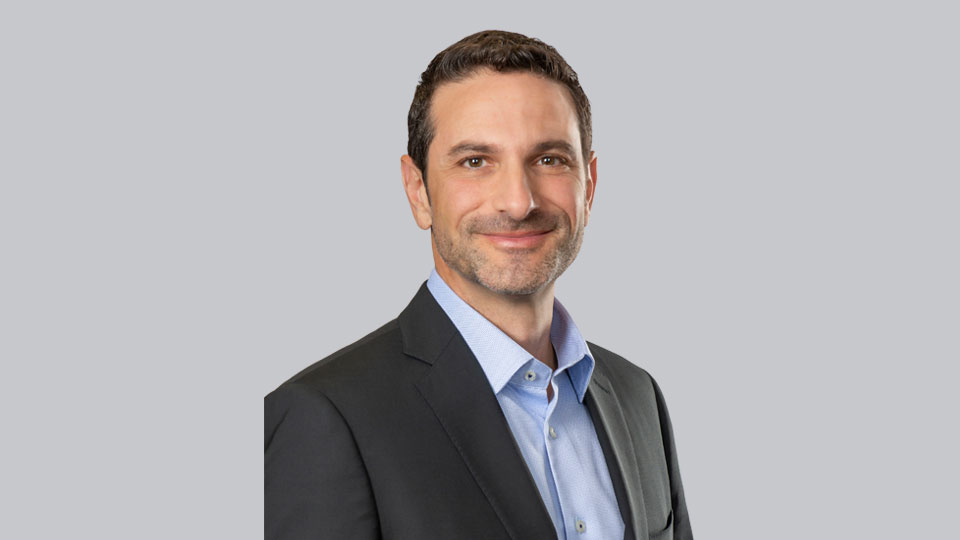 A man is smiling slightly, wearing a dark suit jacket and light blue collared shirt, against a plain, light grey background.