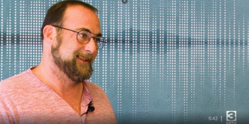 Smiling man in a pink shirt with glasses and short beard against a teal background with vertical dot patterns.