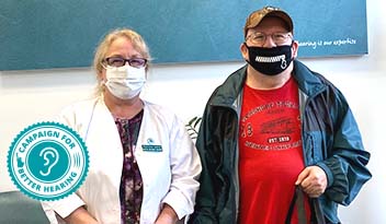 Two masked individuals stand side by side; one dressed in a white lab coat with a name badge and the other in casual attire with a red shirt and cap. A 'Campaign for Better Hearing' logo is displayed in the corner.