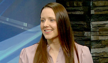 A smiling person with long brown hair wearing a light purple top indoors, framed by a stone wall and blue panels in the background.