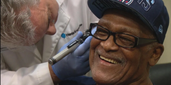 A man smiling while a hearing care professional conducts a hearing evaluation with specialized equipment.