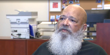 Man with a white beard in an office environment, featuring a blurred bulletin board and printer in the background.