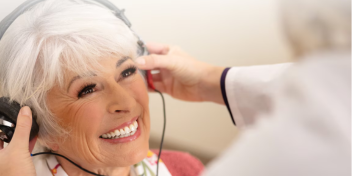 A smiling older woman wears headphones as an audiologist adjusts them during a hearing test in a clinical setting. The interaction highlights personalized care at a hearing clinic. No visible text.