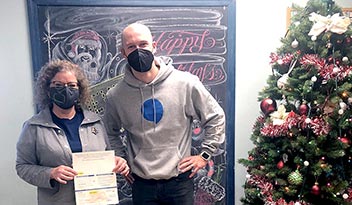 Two individuals wearing masks stand indoors; one holds a certificate. Behind them is a chalkboard featuring a festive Santa drawing with 'Happy Holidays' text, next to a decorated Christmas tree.