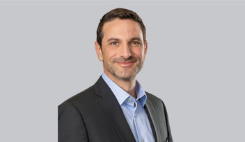 A man with short, dark hair, wearing a suit jacket over a light blue dress shirt, is smiling while facing the camera against a plain gray background.