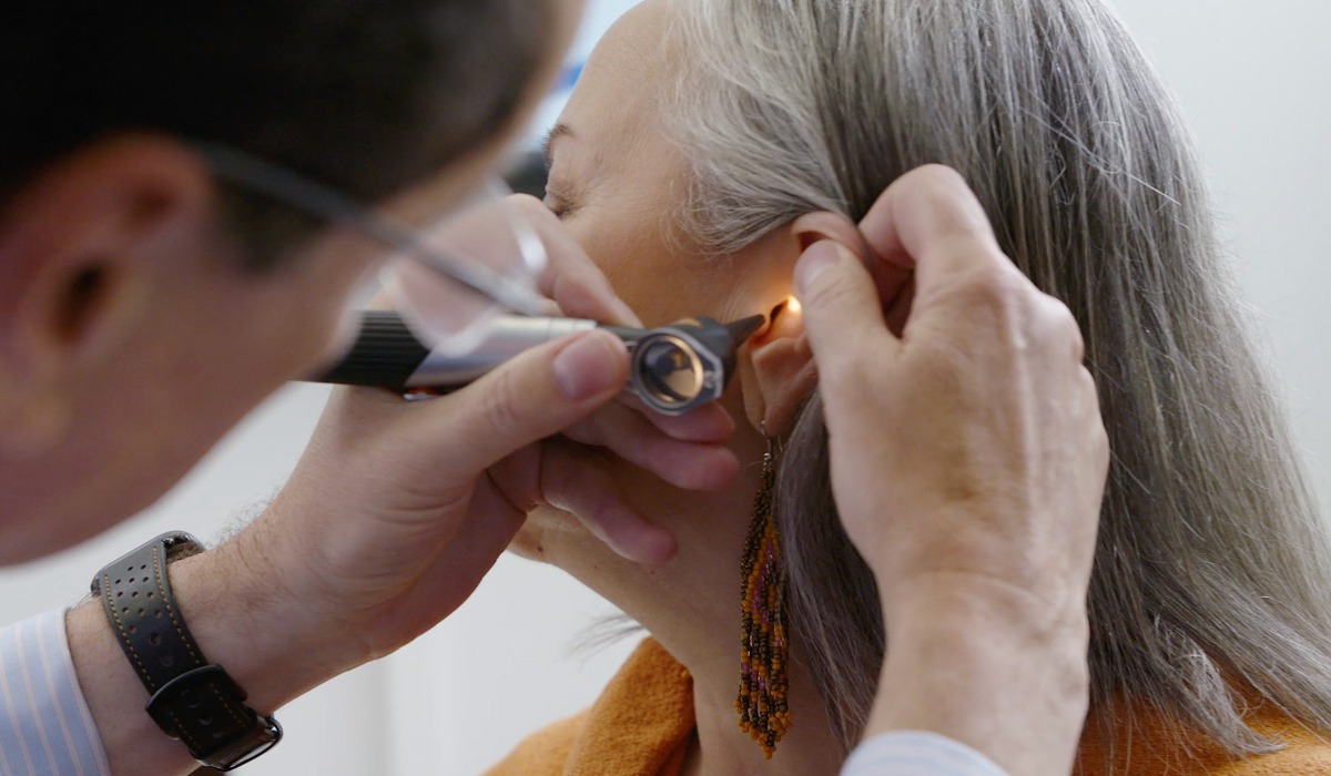 A healthcare professional uses an otoscope to examine the ear canal of a woman with gray hair wearing orange clothing and dangle earrings in a clinical setting.
