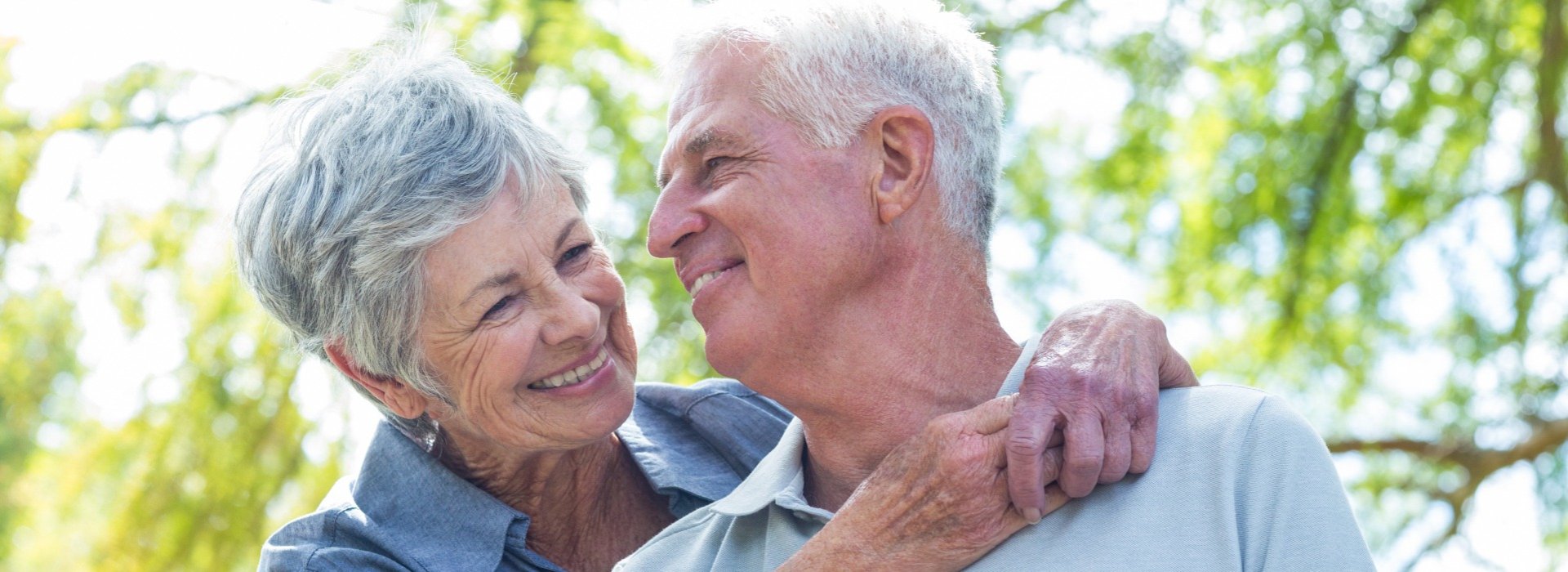 An elderly couple embraces warmly outdoors under bright sunlight, surrounded by a blurred green backdrop of trees. The man's visible ear could imply hearing aid considerations or hearing health awareness.