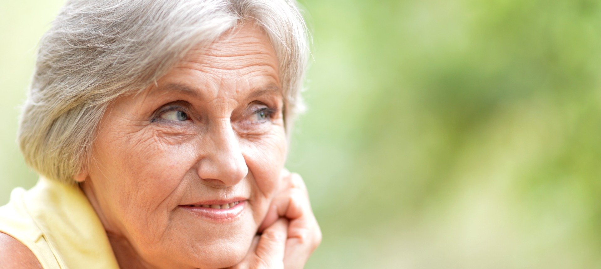 A senior individual with short gray hair wearing a yellow shirt sits outdoors against a blurred green natural background, suggesting a relaxed and peaceful environment. Face obscured for privacy.