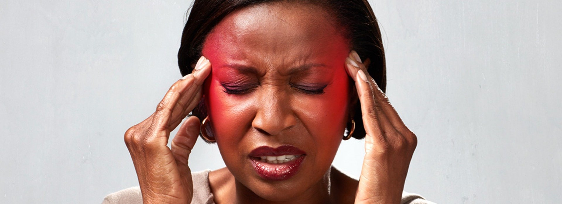 Hands gently press against the sides of a person's head, with a plain, light gray background in a professional setting. The image evokes themes of potential hearing care or discomfort. No text visible.