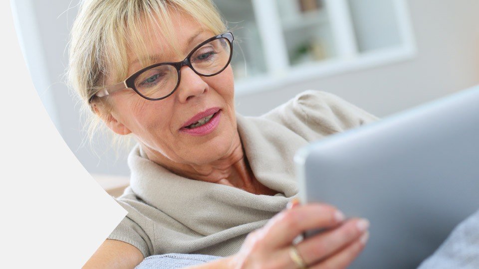 A woman, wearing glasses and a light-colored shirt, is holding and looking at a tablet, sitting in a bright, modern room with shelves in the background.