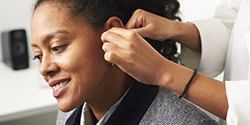 A woman's ear being fitted with a hearing aid by a healthcare professional in a clinical environment.