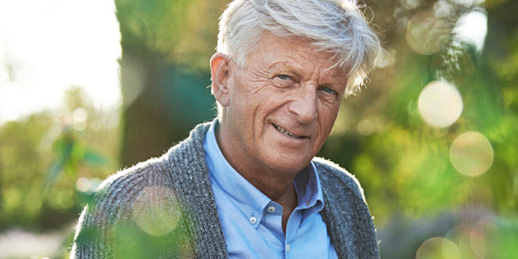 An elderly man smiling warmly in a sunlit garden, wearing a light blue shirt and a gray knitted cardigan.