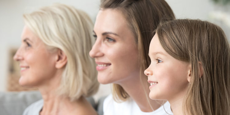 Three females of different ages are sitting side by side, smiling and looking off to the right in a relaxed indoor setting.