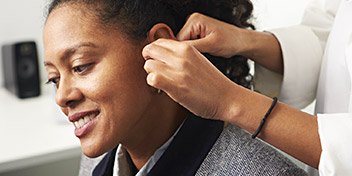 A woman's ear is being fitted with a hearing aid by a healthcare professional in a clinical setting.