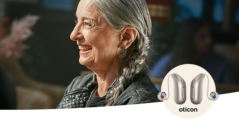 An older individual with braided gray hair is wearing an Oticon hearing aid discreetly behind the ear. The context is a cozy, indoor setting with blurred surroundings. An inset displays two silver Oticon hearing aid models, emphasizing their sleek design and functionality for advanced hearing care.